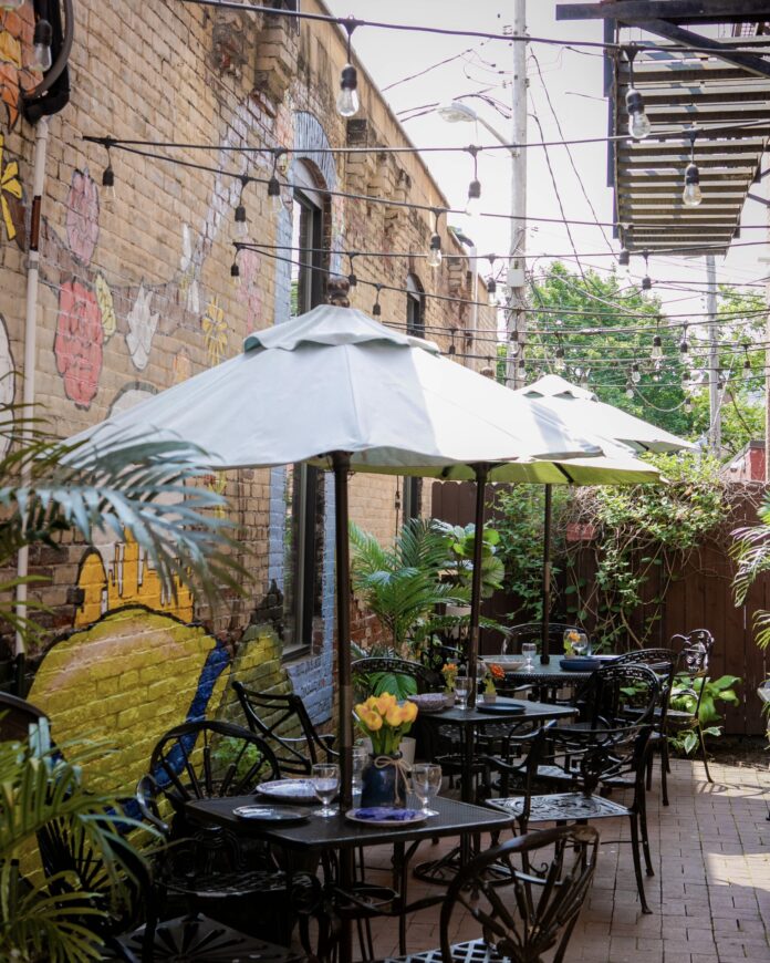 a narrow passage between brick buildings with graffiti set up for outdoor dining with black iron tables and chairs and white cafe umbrellas