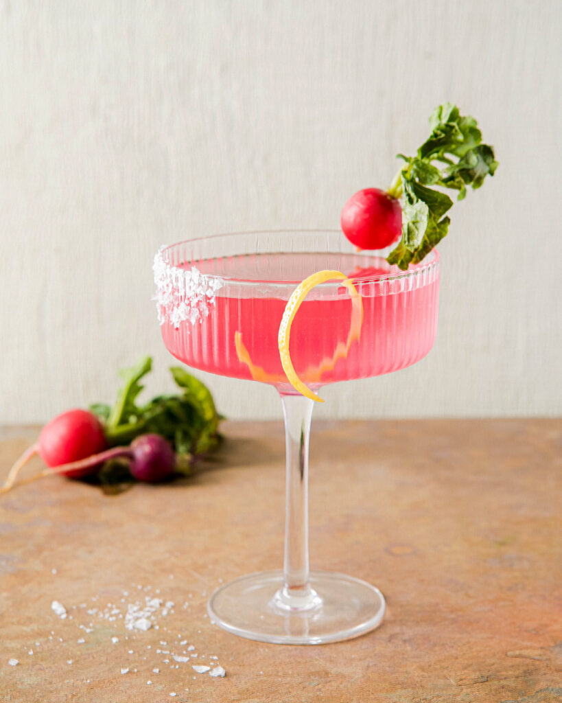 Pickled Radish Martini sits in a martini glass, light pink in color, with a lemon peel and radish acting as a garnish.