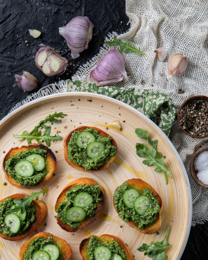 green nasturtium pesto on crostini with small cucumber slices on a beige tray garnished with arugula, a black background, green floral fabric, tan burlap and fresh garlic with purple skins
