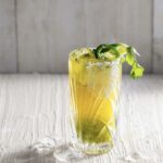 a yellow and green cocktail made of verde and beer in a clear glass garnished with jalapeño and cilantro with a white background
