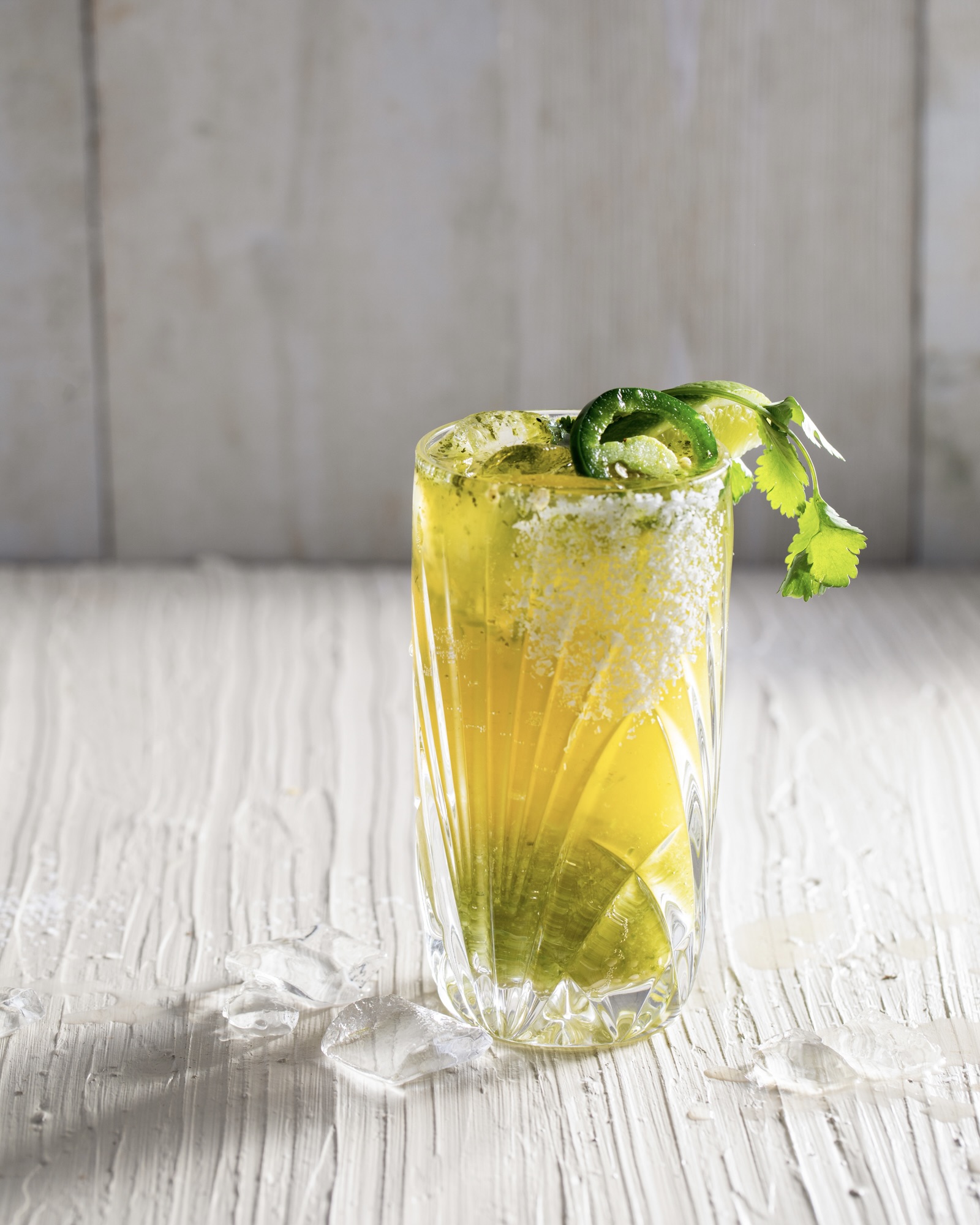 a yellow and green cocktail made of verde and beer in a clear glass garnished with jalapeño and cilantro with a white background