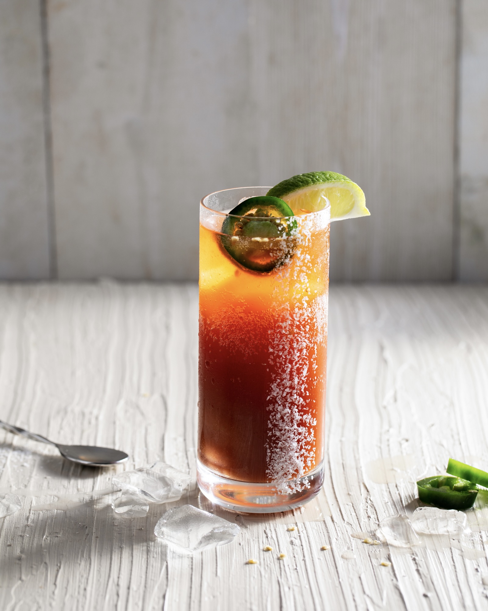 a look in photo of a tall glass with Bloody Mary mix and beer, called a rogue michelada, with a white back ground and lime wedges