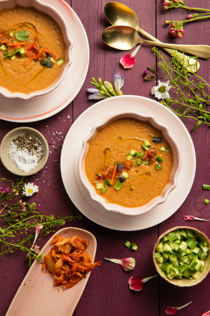An aerial view of two bowls of Kimchi Melon Gazpacho, orange in color, sitting on wooden slates purple in color and surrounded by flowers and herbs. Kimchi Melon Gazpacho Recipe