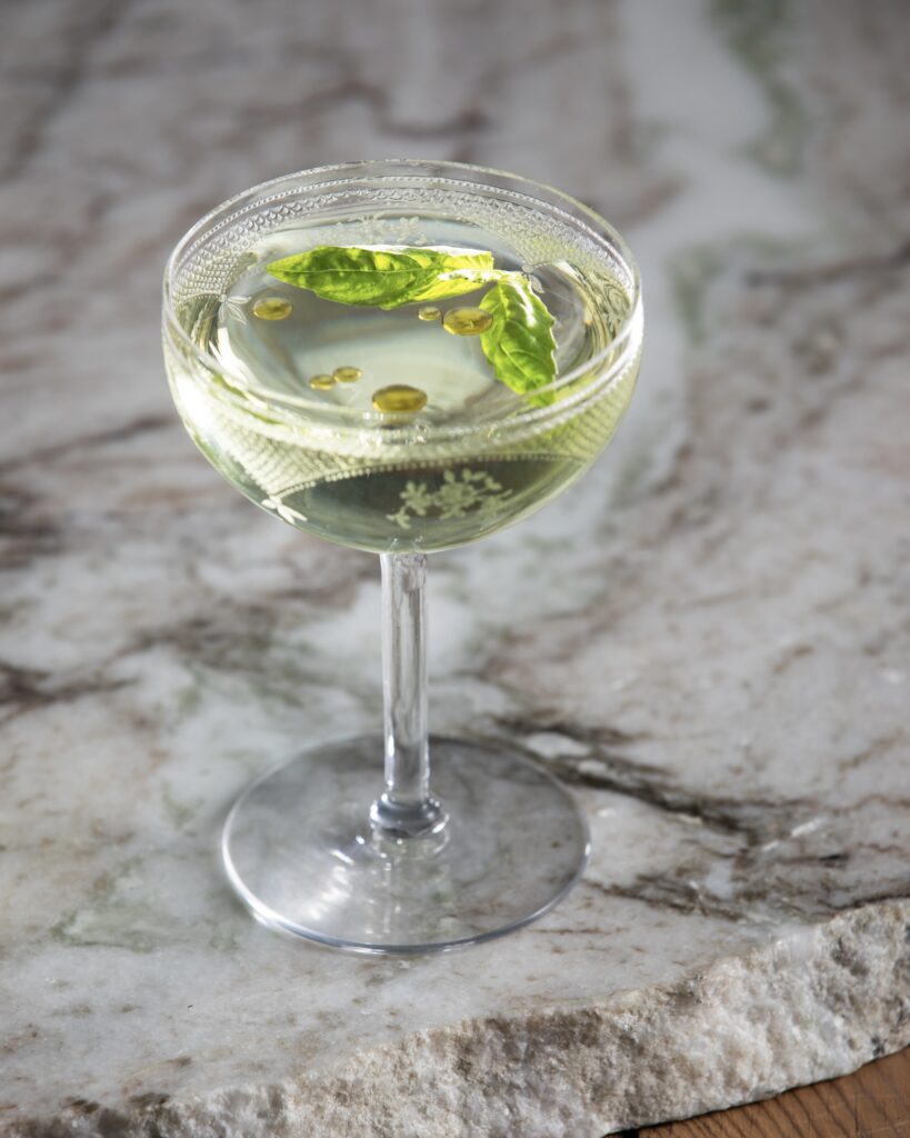 a lemon basil martini cocktail in a coupe glass sitting on a white and grey marble slab with fresh basil and olive oil floating on the top of the cocktail