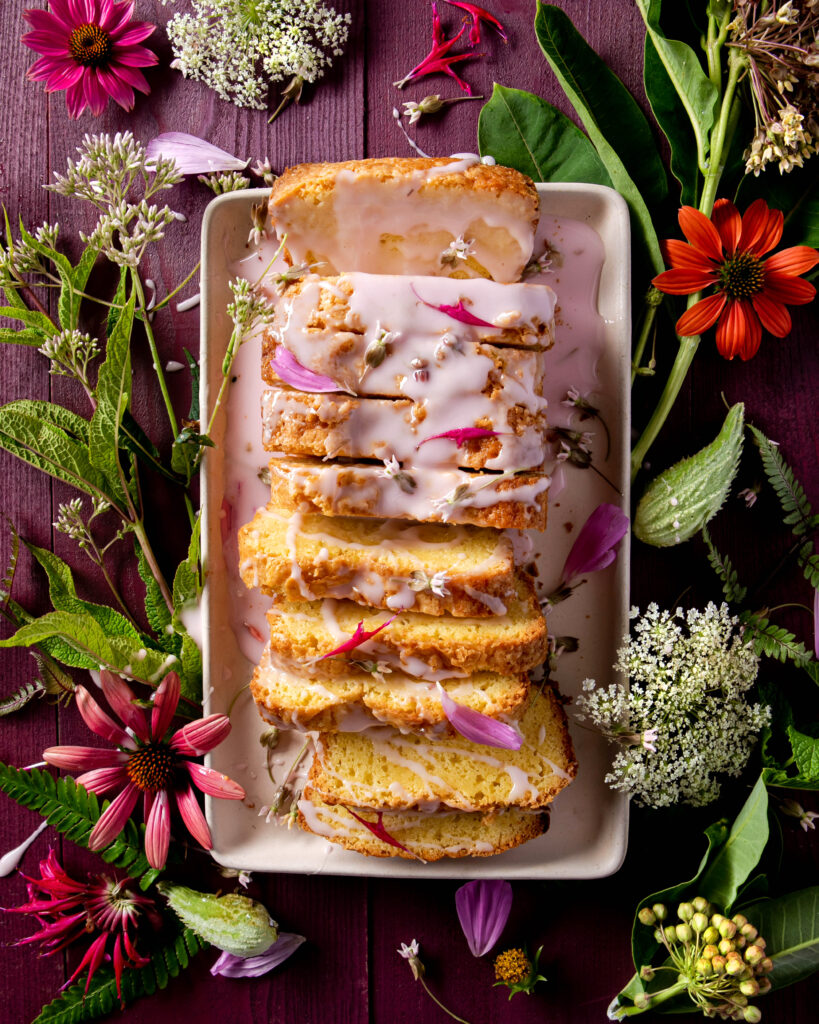 An aerial view of Milkweed Flower Pound Cake sitting sliced on a white serving tray, surrounded by flowers. Milkweed Flower Pound Cake Recipe