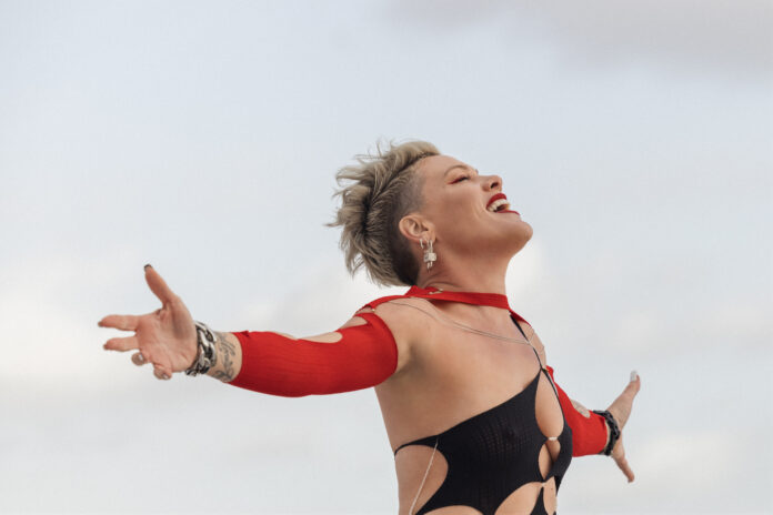 A white woman, music artist pink, holds her arms out to to sides and tilts her head to the sky