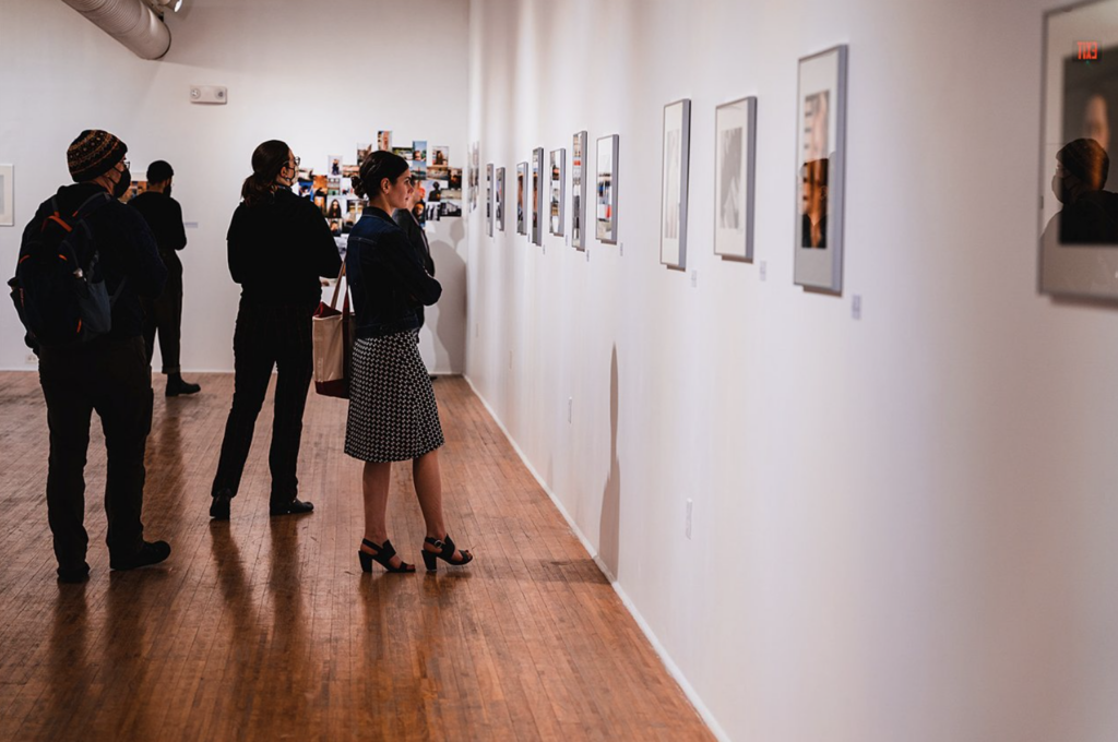 A group of onlookers take in framed artwork on a white gallery wall.