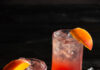 So What Cocktail, Inspired by P!nk a look in at 2 identical dark peachy pink cocktail on a black background with ice cube and peach wedges as garnish
