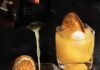 Sunflower Cocktail, Inspired by Post Malone 2 orange cocktail in rocks glasses on a black surface with dehydrated orange slices as garnish. The front cocktail is being poured by a woman's hand