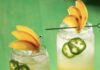 Man, I Feel Like a Woman Cocktail Inspired by Shania Twain 2 glasses with peach colored cocktails garnished with peach slice fans and jalapeño slices sitting on a vibrant green wooden surface