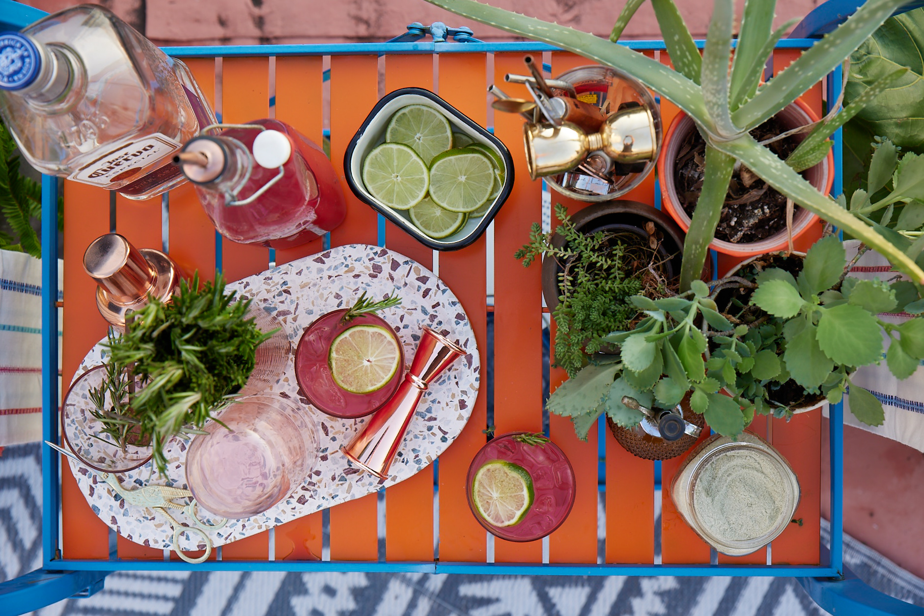 an arial shot of an orange slatted bar cart with a premixed cocktail a bottle of tequila, lime and herb garnish and several smaller potted plants