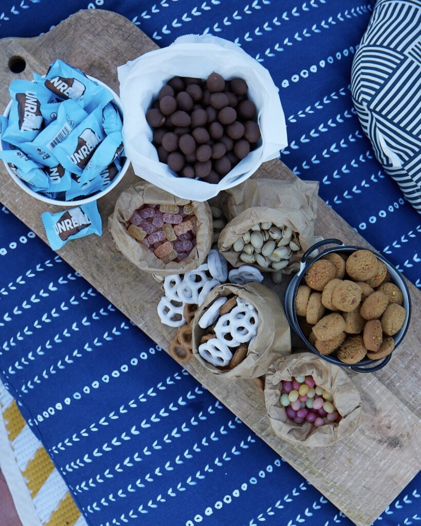 wrapped candies, chocolate covered nuts, yogurt covered pretzels, mini sandies cookies, pistachios in the shell, and jelly beans on a paper runner on a blue tablecloth