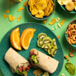 White Bean Tuna Salad Wraps sit on a green plate with two orange slices, and a few pieces of kiwi.