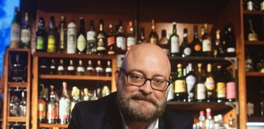 A white male stands behind a bar surrounded by bottles and mixed drinks.