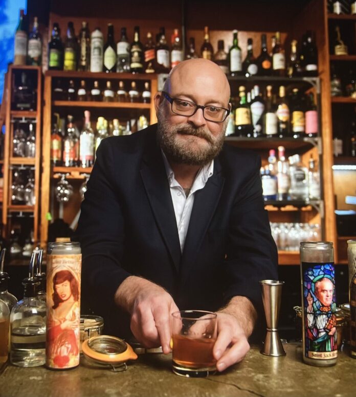 A white male stands behind a bar surrounded by bottles and mixed drinks.