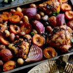 A sheet pan with chicken thighs, halved apricots, purple onions and baby potatoes with a stack of creamy yellow plates and gold forks. Asian Stone Fruit Chicken Bake Recipe