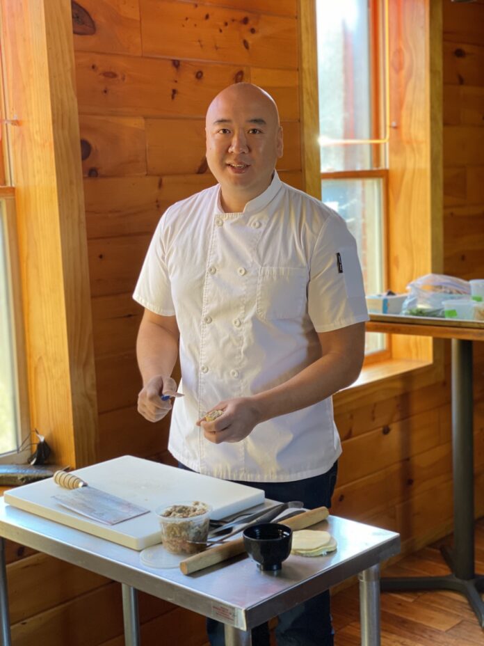 Chef Roger Li, in a white chefs jacket, stands facing the camera over a small prep table.