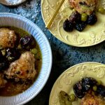 An arial view of a round white baking dish with chicken thighs, olives, prunes, and capers, and two yellow dishes with single servings of the same with gold forks