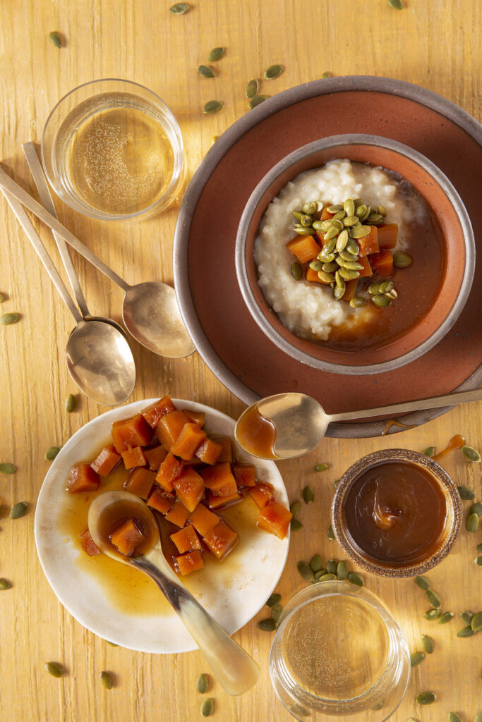 Rice Pudding with Candied Pumpkin and Caramel sits in an orange and brown bowl. Bowls of candied pumpkin, and caramel sit below the pudding. Rice Pudding recipe