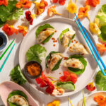 An aerial view of Pork Dumplings with Edible Flowers on a white plate. Blue chopsticks sit to the right side of the plate. Edible Flowers sit around the plate.