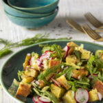 Three individual blue bowls above a green serving bowl filled with a vibrant potato salad of yellow, green, red, and purple ingredient
