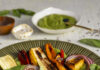 Nut and Dairy Free Pesto and Summer Vegetables A green plate with red, yellow, green, orange, and purple grilled vegetables garnished with fresh basil and a small white bowl of pesto on the side.