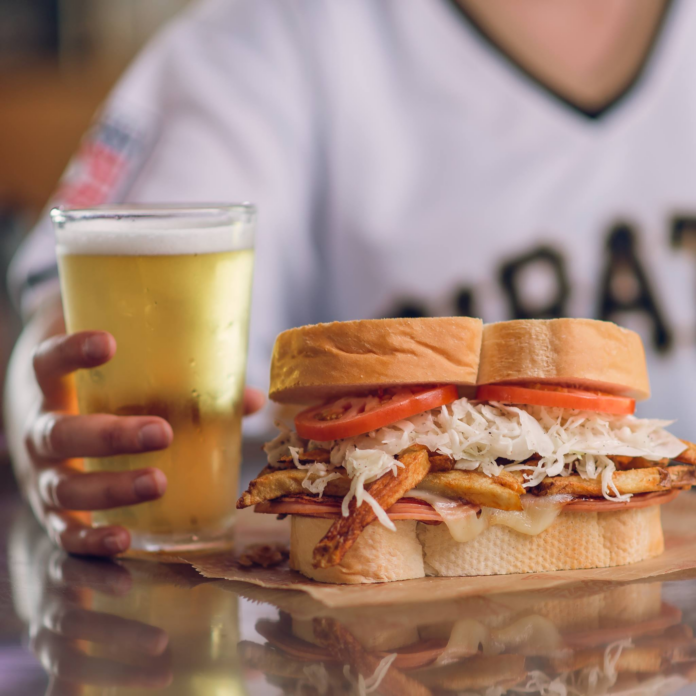 A white male's hand is wrapped around a beer. Sitting next to the drink is a Primanti's sandwich.