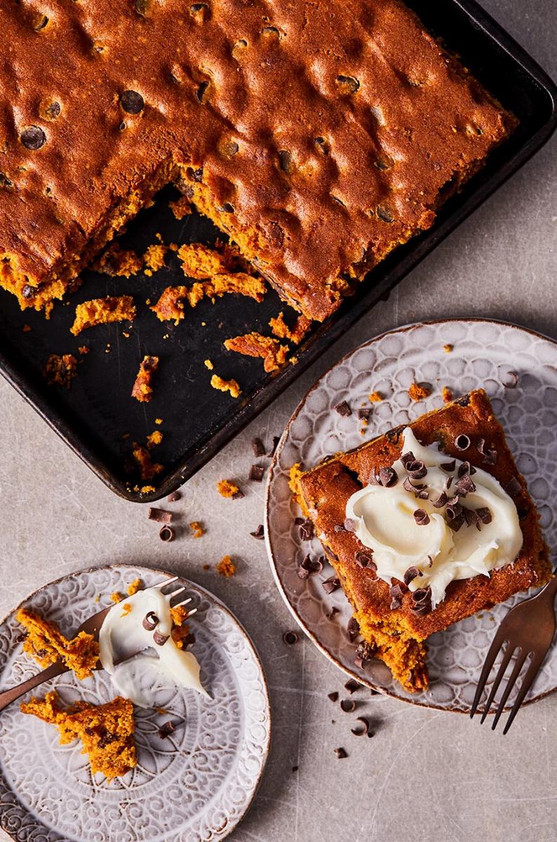 A baking pan of pumpkin blondies with a square slice topped with cream cheese frosting and chocolate shavings on a small plate.