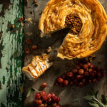 Flat-lay image of a savory cheesecake, roasted grapes on a weathered table.