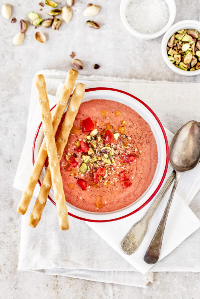 An above shot of Tomato Strawberry Gazpacho, pink-red in color, served in a bowl with three thin breadsticks on the side.