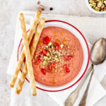 An above shot of Tomato Strawberry Gazpacho, pink-red in color, served in a bowl with three thin breadsticks on the side.