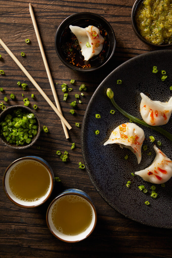 A spread of dumplings and Ginger and Scallion Sauce on a wooden table. Chopsticks sit to the left of the dumplings. Ginger and Scallion Sauce Recipe