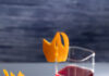 Forever Young Cocktail, Inspired by Rod Stewart Two layered cocktails, orange on the bottom and red on the top, in clear square glasses with citrus flames as garnish and 2 lemon wedges to the right of the glasses.