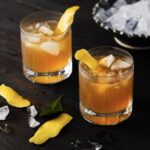 2 identical whisky cocktails in rocks glasses, garnished with lemon peel with a black background
