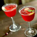 Two cocktails, bright ref in color sit in wine glasses and are garnished with lemon round, pomegranate arils, and mint leaves