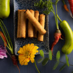Flat-lay image of a plate of spring rolls surrounded by colorful vegetables.