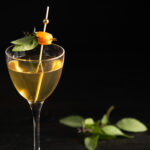 A pale yellow cocktail in a wine glass garnished with basil
