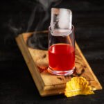 A cocktail red in color sits on a wooden board that is slightly burnt with smoke pouring out. Coo Coo for Coconuts Negroni