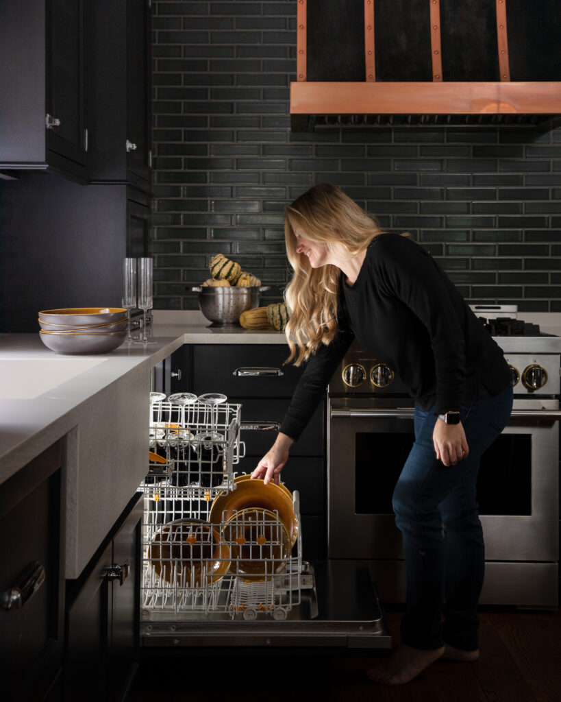 a woman in a black long sleeve and jeans reaches into an open dishwasher