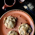 Three large Snickers Levain Bakery cookies on a plate with a cup of coffee, a small vessel of creamer, and raw sugar on a spoon