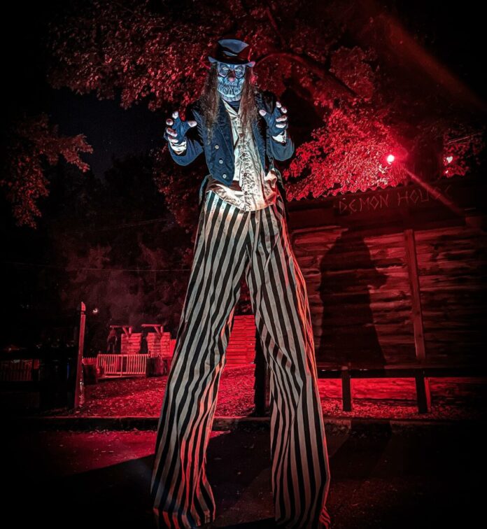 haunted attractions A clown in striped pants on stilts stands outside in front of red lights.