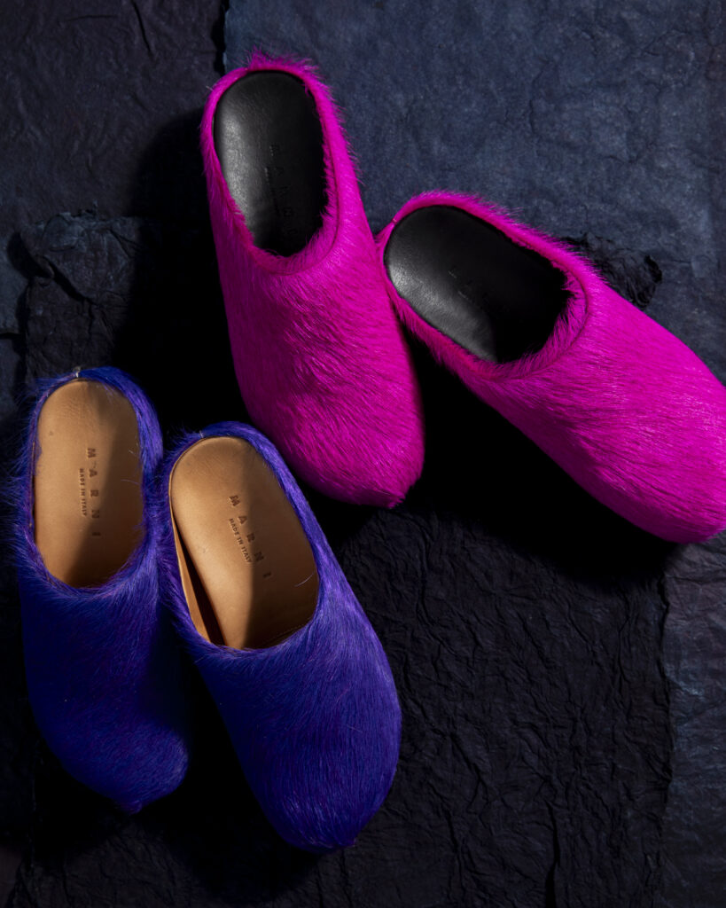 Pink calf hair women's mules and purple calf hair men's mules on a black backdrop.