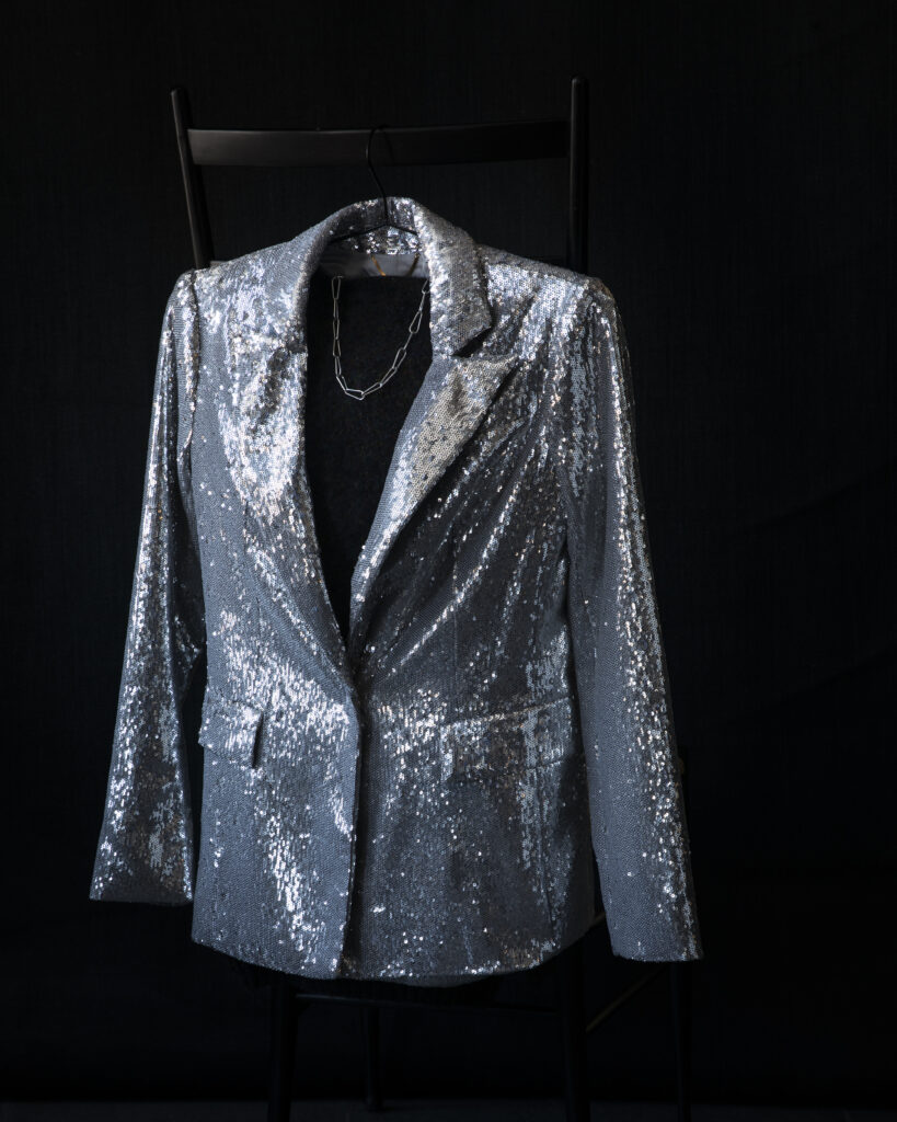 A silver blazer is displayed on a dark background, styled with a black shirt and necklace.