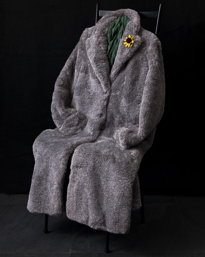 A grey fur coat with a sunflower on the collar laid out on a black background in a sitting position.