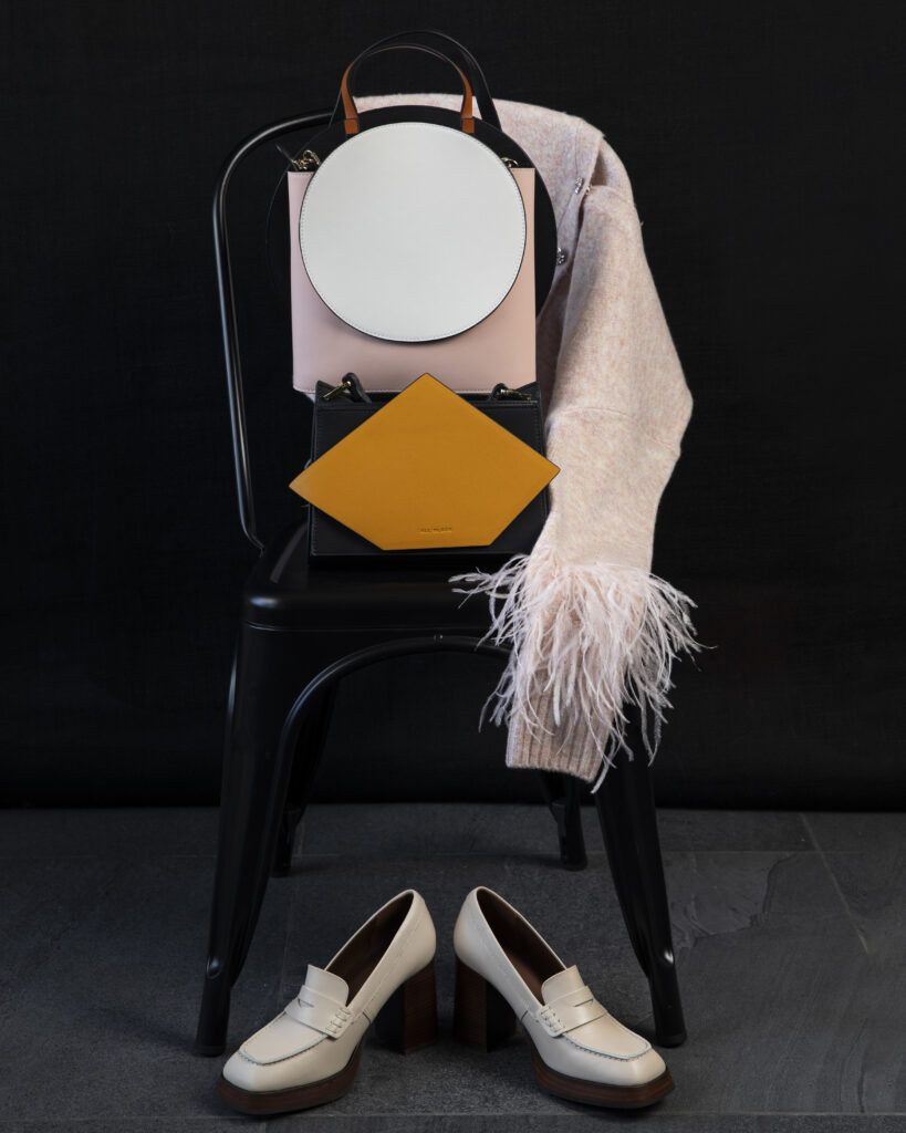 Two geometric purses modeled with a pin sweater and white shoes on a chair.