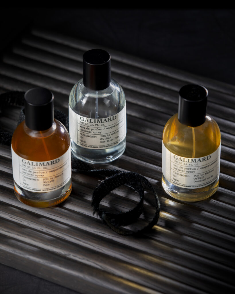 Three bottles of colorful perfume sit on a black stripped tabletop.