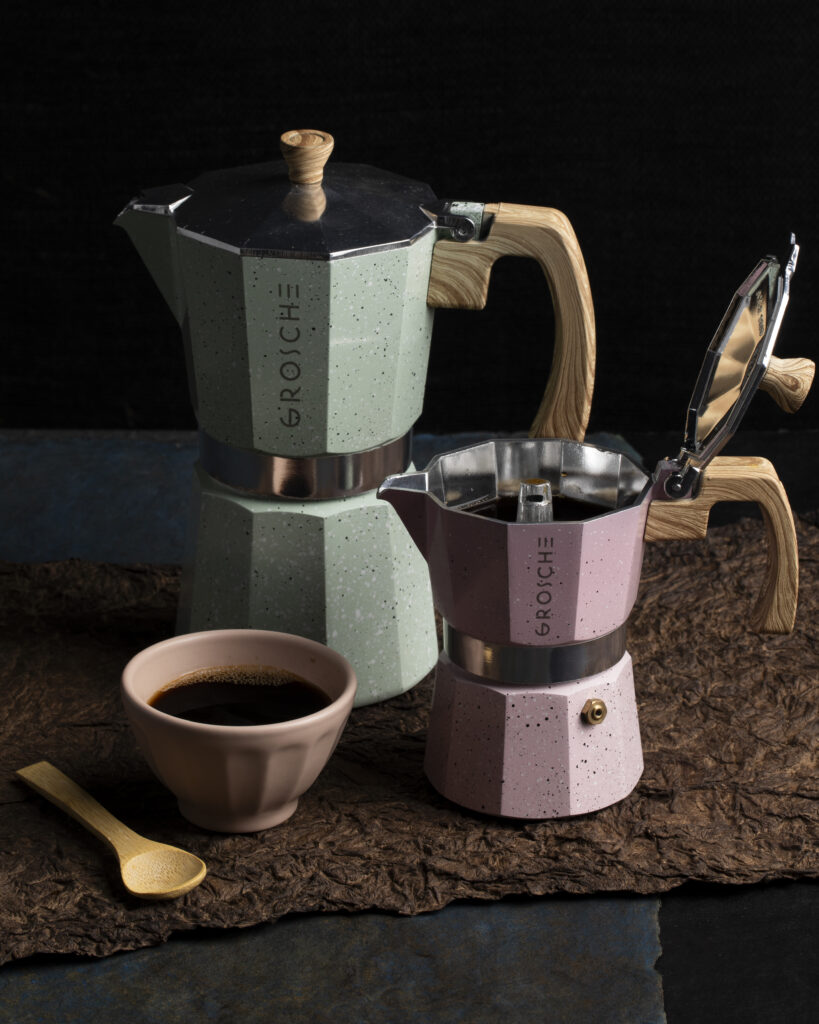 Two coffee moka pots, one light blue and one pink beside a bowl of coffee grounds and a spoon.