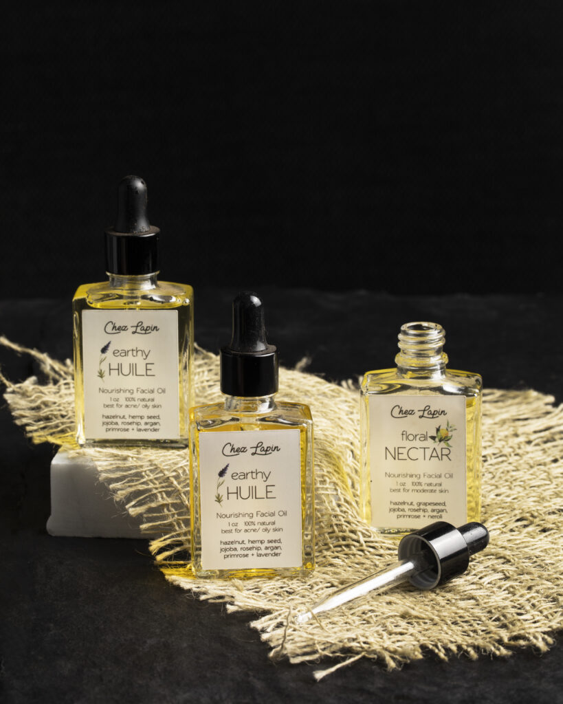 Three facial oils are laid out on a fringe beige rug.
