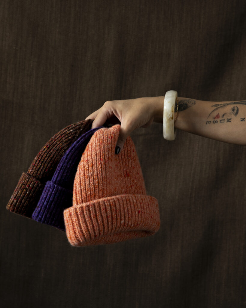 A variety of orange, brown, and dark blue beanies held by a hand with a bracelet on.
