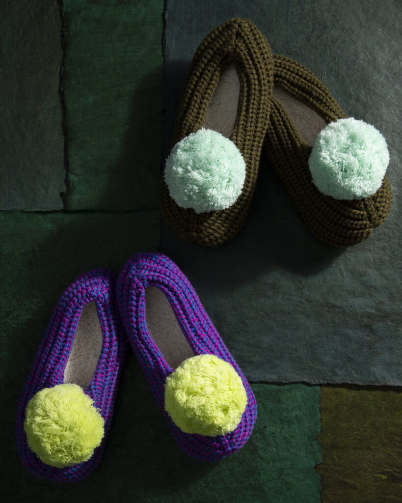 Two pairs of knitted pom pom shoes on a dark green wood background.